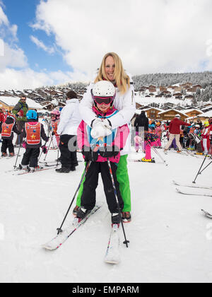 Mother and small child in skiing clothes on skis  prior to a skiing lesson with slopes  and chalets behind Stock Photo