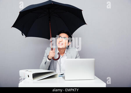 Happy asian businessman sitting at the table with laptop and holding umbrella over gray background Stock Photo
