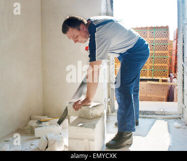 middle-aged man worker in blue trousers and gray t-shirt saws silicate brick in the room of new house Stock Photo