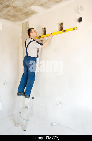 middle-aged man worker in blue trousers and gray t-shirt checks the wall with a level in the room of new house Stock Photo