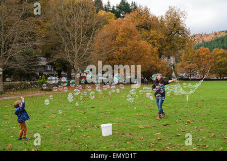 A young boy plays with lots of small soap bubbles made by a bubble wand on village  green in Betws-y-Coed Conwy North Wales UK