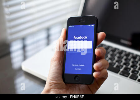 Hand holding Iphone with mobile application for Facebook on the screen, laptop on background Stock Photo