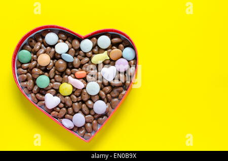 Heart shaped box filled with small chocolates balls with a lot of them scattered on the yellow background Stock Photo