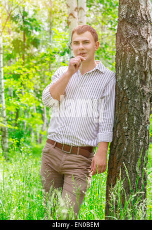 Romantic guy in the park, standing near birch tree with straw in the mouth, carefree weekend, enjoying beauty of spring nature Stock Photo