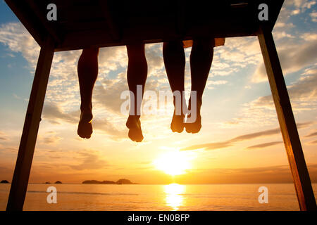 silhouette of feet of couple sitting on the pier at sunset beach, low angle view