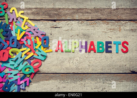 The colorful words 'ALPHABETS' made with wooden letters next to a pile of other letters over old wooden board. Stock Photo