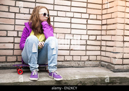 Blond teenage girl in a sunglasses sits on her skateboard near urban brick wall, photo with retro tonal correction, instagram ol Stock Photo