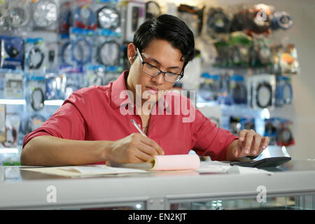 Young asian entrepreneur owning a computer shop and checking bills and invoice with calculator, doing budget and reviewing taxes Stock Photo