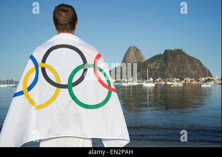 RIO DE JANEIRO, BRAZIL - MARCH 24, 2015: Man draped in Olympic flag stands on the shore of Botafogo Bay in front of view of Suga Stock Photo