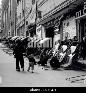 1950s historical picture, Hong Kong, on a sloping side-street, a row of pulled rickshaws or passenger carts. Most of the rickshaws have their hoods up, but three have their hoods down showing they are available for hire. Their operators are playing a game on the street. Rickshaws have been a popular form of taxi transport for centuries, their name coming from the japanes for human-powered transport. Offices of the South China Morning Post newspaper can be seen, and the rickshaws are parked outside the Blue Theatre and nightclub. Stock Photo