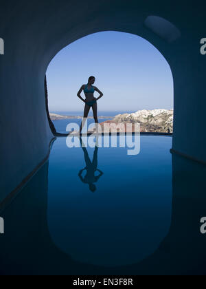 Silhouette of woman in bikini standing at edge of infinity pool with arch and rock formation Stock Photo