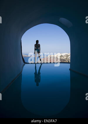 Silhouette rear view of woman in bikini standing at edge of infinity pool with arch and rock formation Stock Photo