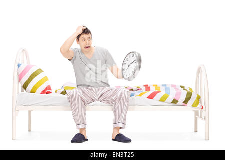 Shocked young man sitting on a bed in his pajamas and looking at the time isolated on white background Stock Photo