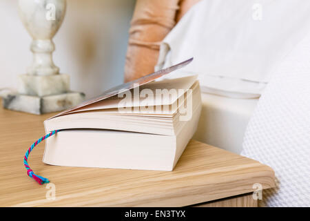 Everyday scene of a closed paperback book reading matter with bookmark marking a page on a bedside table at side of a bed. England UK Britain Stock Photo