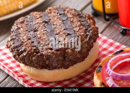 A juicy barbecued hamburger with grill marks on a rustic picnic table with tomato, onion, mustard, ketchup, and corn. Stock Photo