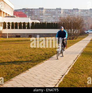 elderly man wearing glasses, a blue jacket, black trousers and a gray hat riding a bicycle on the sidewalk of concrete tiles Stock Photo