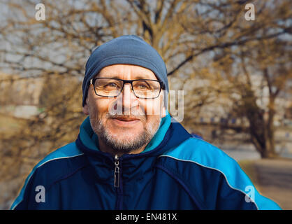 portrait of a smiling elderly man wearing glasses, a gray hat and a blue jacket in the city park Stock Photo