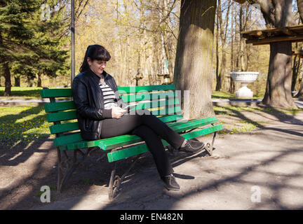 young beautiful woman in a black leather jacket and black pants sitting on a park bench holding a tablet Stock Photo