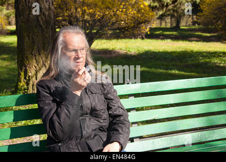 elderly gray-haired man in glasses, black jacket smoking a cigarette sitting on a bench in city park on sunny spring day Stock Photo