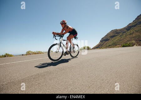 Image of young woman cycling on the country road. Fit female athlete riding down hill on bicycle. Woman doing cycling training. Stock Photo