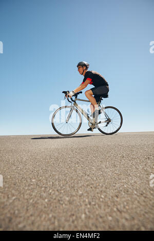 Side view of male cyclist riding bicycle on flat road against blue sky. Low angle view of caucasian triathlete training for comp Stock Photo