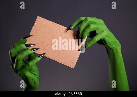 Green hands with long black nails holding empty piece of cardboard, Halloween theme Stock Photo