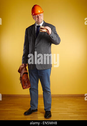 Male engineer in room, he eats sandwich and holds old leather briefcase, parquet floor and yellow wall Stock Photo