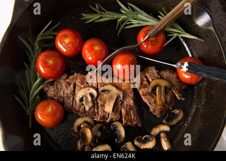 Grilling Strip Loin Steak in Cast Iron Frying Pan: Steak  is cooked  and sliced  - shown with grilled tomatoes and mushrooms Stock Photo