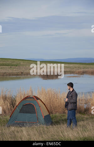 A young adult male standing next to a tent drinking coffee while on a camping trip. Stock Photo