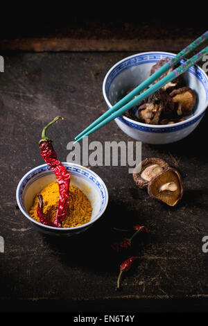 Tumeric powder and shiitake mushrooms in porcelain bowls, served with red hot chili peppers and turquoise chopsticks over dark t Stock Photo