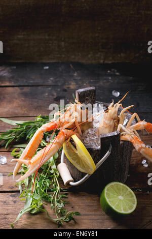 Raw langoustines on ice with herbs and lemon served in wooden bucket over wooden table Stock Photo