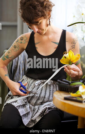 A tattooed woman florist cuts the white ribbon on a boutonniere made of a yellow orchid with a pair of blue handled scissors. Stock Photo