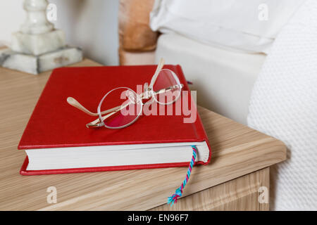 Generic plain red hardback book and reading glasses on a bedside table at the side of a bed in a domestic bedroom at home. England, UK, Britain Stock Photo