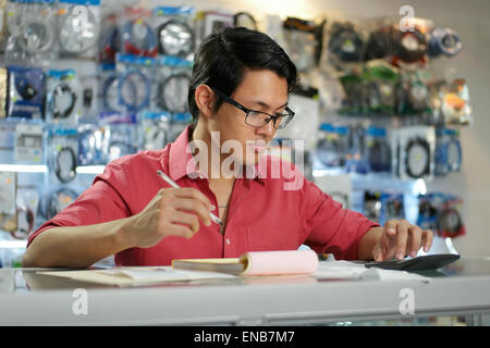 Young asian entrepreneur owning a computer shop and checking bills and invoice with calculator, doing budget and reviewing expen Stock Photo