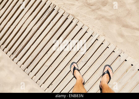 Legs of Young Caucasian man standing on wooden footbridge over a sandy beach Stock Photo