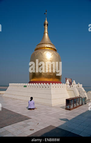 A Burmese woman kneels in prayer in front of the golden Bupaya stupa against a cloudless blue sky Stock Photo