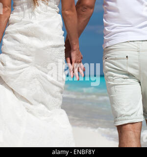 Young beautiful newlyweds in love holding hands and walking away on tropical sandy beach, back view Stock Photo