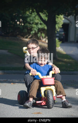 Two brothers, age seven and four ride a big wheel three wheeler in their driveway. Stock Photo