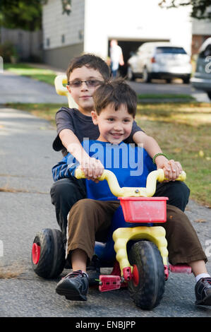 Two brothers, age seven and four ride a big wheel three wheeler in their driveway. Stock Photo