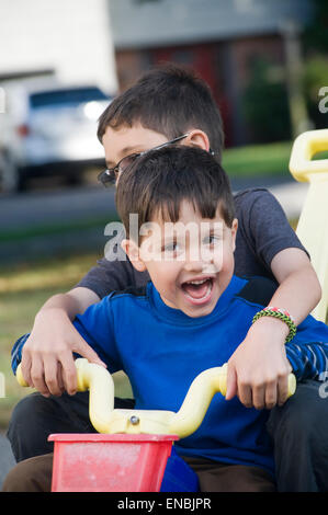Two brothers, age seven and four ride a big wheel three wheeler in their driveway. Stock Photo