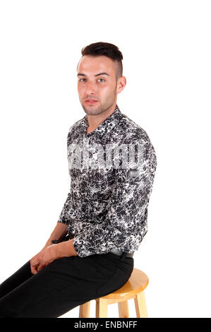 A young handsome man sitting in profile in black jeans and black and white shirt, isolated on white background. Stock Photo