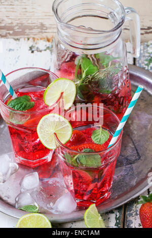 Two glasses with retro cocktail tubes and glass jug of homemade strawberry lemonade, served with fresh strawberries, mint, lime Stock Photo