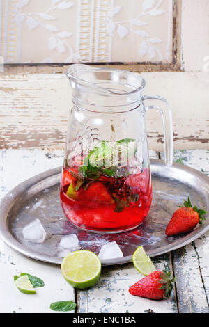 Glass jug of homemade strawberry lemonade, served with fresh strawberries, mint, lime and ice cubes over old white wooden table. Stock Photo