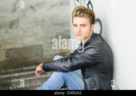 Attractive young man wearing black leather jacket and jeans sitting on stairs Stock Photo