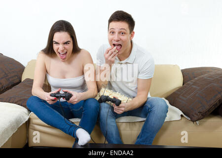 young smiling couple in love on sofa sitting and playing video games in livingroom at home with popcorn. Man and woman having fu Stock Photo