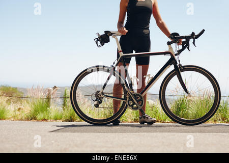 Low section shot of female athlete standing with her bicycle. Woman cyclist with her bike on country road. Cropped shot of femal Stock Photo