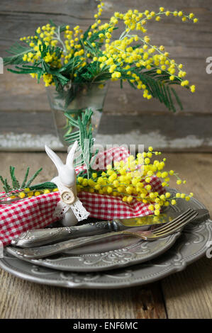Spring festive dining table setting with yellow mimosa flowers, candles, napkins and vintage cutlery on a wooden board Stock Photo