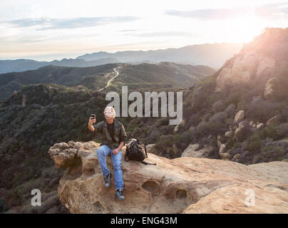 Older Caucasian man taking cell phone photograph on rocky hilltop Stock Photo