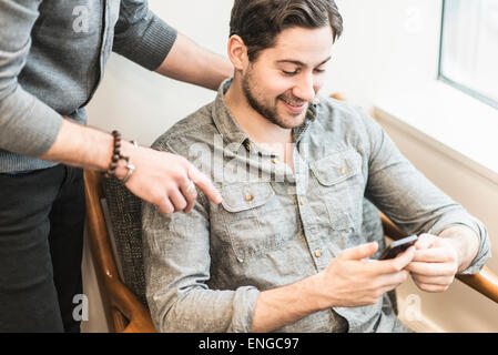 A man seated checking his smart phone and a colleague looking over his shoulder. Stock Photo