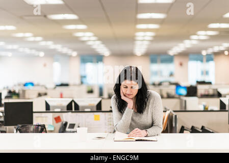 A woman seated with her hand resting on her chin, reading a book at a desk. Stock Photo